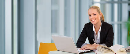 Junge Frau in Anzug sitzt an einem Tisch mit Laptop und Buch, lächelt in einem modernen Büro.