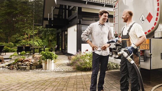 Zwei Männer im Gespräch neben einem Lieferwagen auf einem gepflasterten Außenbereich eines modernen Hauses mit Garten.