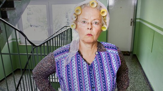 Ältere Frau mit Haarwicklern und schockiertem Gesichtsausdruck steht in einer grünen Treppe.