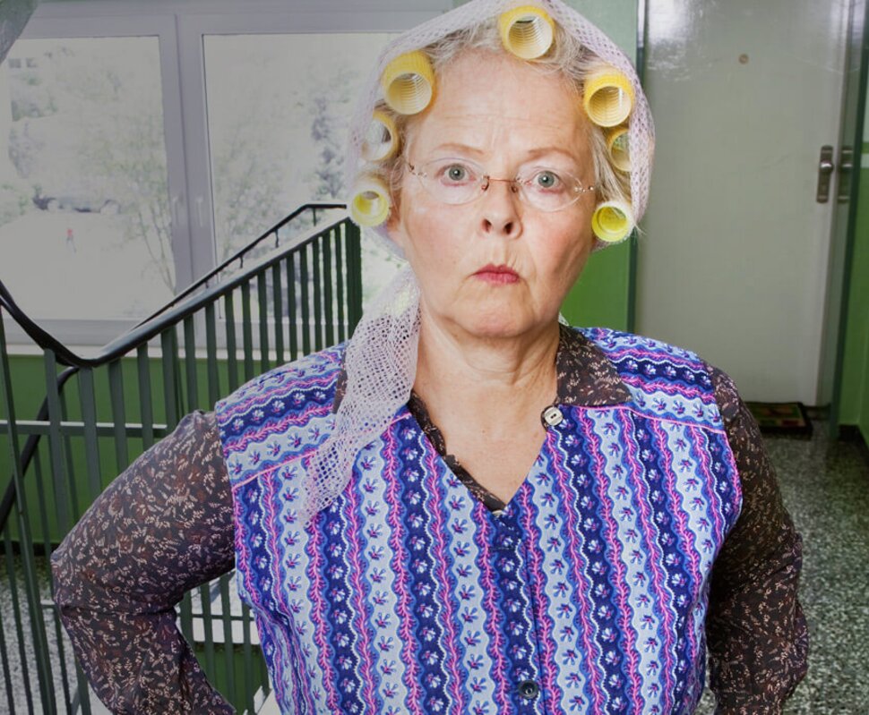 Ältere Frau mit Haarwicklern und schockiertem Gesichtsausdruck steht in einer grünen Treppe.