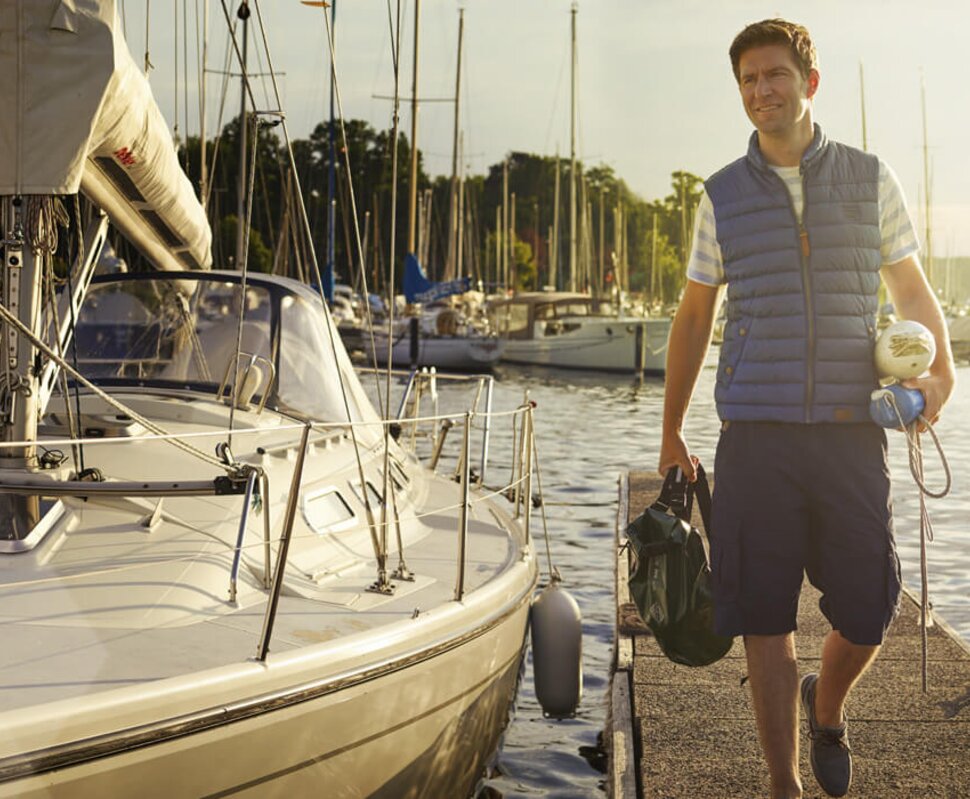Mann mit einem Rucksack und einer Jacke steht an einem Yachthafen neben einem Segelboot. Sonne scheint am Abend.