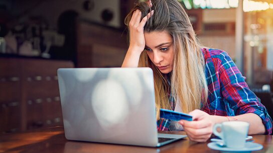 Eine frustrierte junge Frau sitzt an einem Tisch und schaut auf ihren Laptop, während sie eine Kreditkarte in der Hand hält.