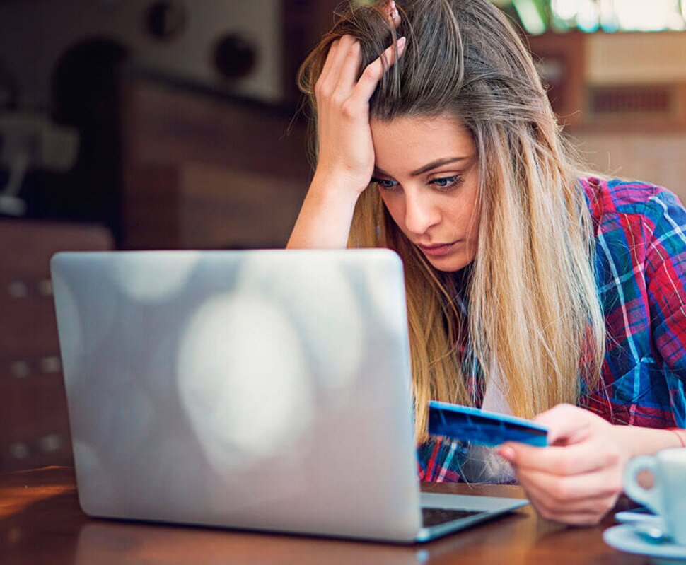 Eine frustrierte junge Frau sitzt an einem Tisch und schaut auf ihren Laptop, während sie eine Kreditkarte in der Hand hält.