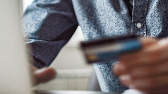 Mann mit Brille und Hemd hält eine Kreditkarte in der Hand und schaut auf einen Laptop-Bildschirm.