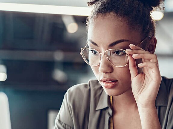 Eine junge Frau mit lockigem Haar trägt Brillen und schaut konzentriert auf einen Laptop, daneben eine Kaffeetasse.