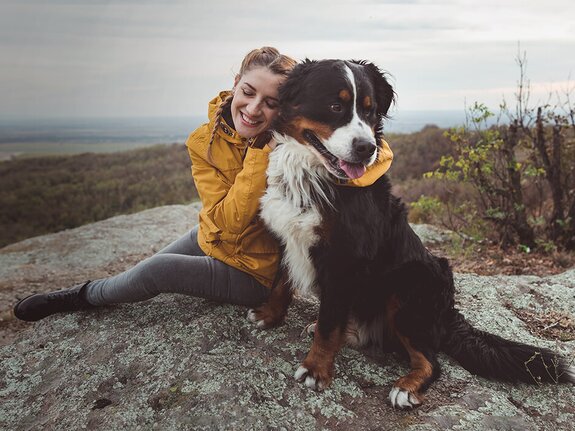 Lächelnde Person in Gelb umarmt einen schwarzen und weißen Hund auf einem Felsen mit Blick auf eine bewaldete Landschaft.