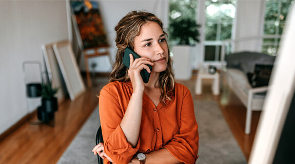 Junge Frau mit längeren Haaren im orangefarbenen Hemd spricht am Telefon in einem modernen Wohnraum.