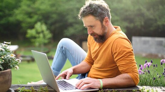 Mann mit Bart sitzt entspannt im Freien und arbeitet an einem Laptop, umgeben von blühenden Blumen und Grün.