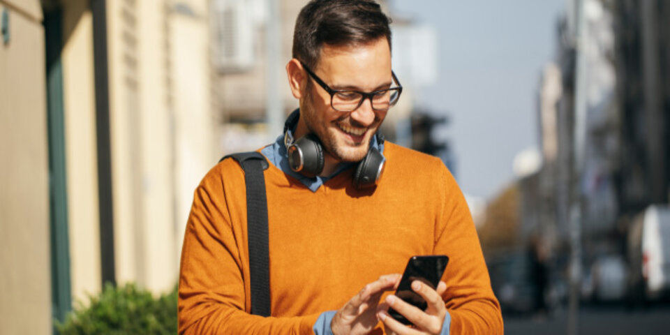 Mann mit Brille und Kopfhörern in orangefarbenem Pullover, der lächelnd auf sein Smartphone schaut.