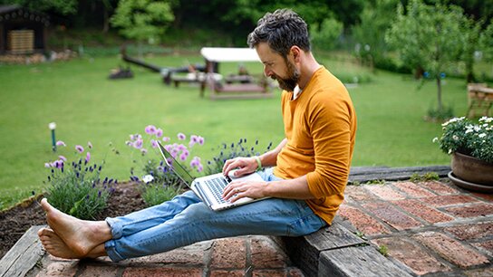 Ein Mann mit Bart sitzt barfuß auf einer Steinmauer und arbeitet konzentriert an einem Laptop in einem Garten.