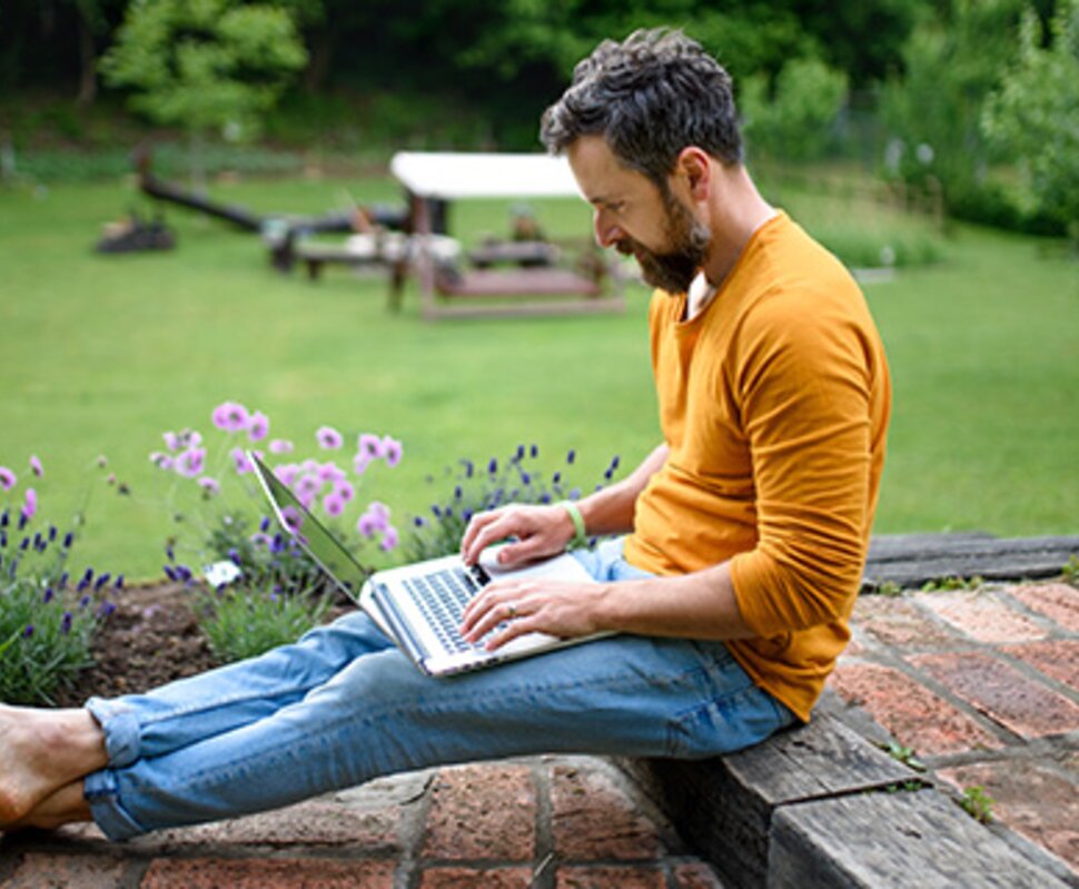 Ein Mann mit Bart sitzt barfuß auf einer Steinmauer und arbeitet konzentriert an einem Laptop in einem Garten.