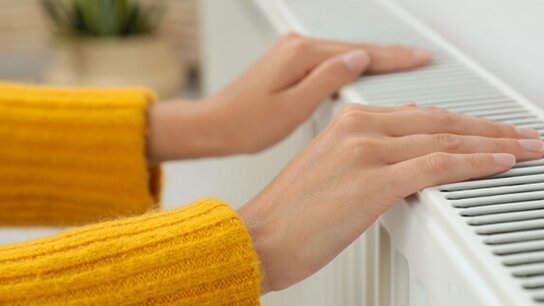 Hände liegen auf einem Heizkörper, während eine Person in einem gelben Pullover die Wärme spürt.