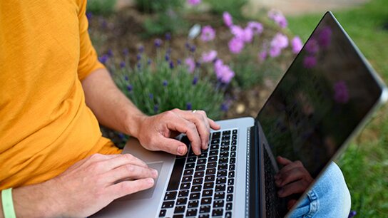 Junger Mann tippt auf einem Laptop, umgeben von lila Blumen und grünem Gras.