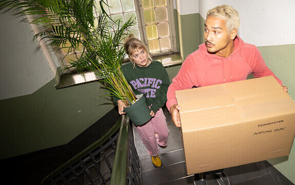 Junge Menschen tragen eine Pflanze und einen Karton die Treppe hinauf, umgeben von natürlichem Licht und einer neutralen Wandfarbe.