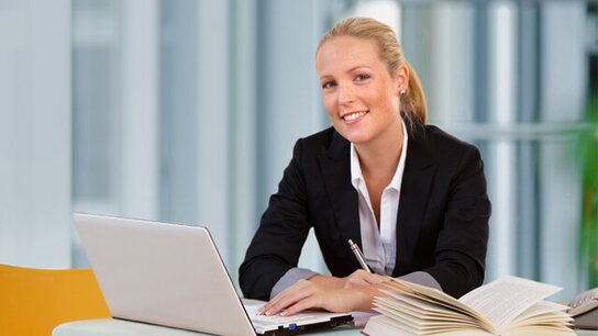 Junge Frau in Anzug sitzt an einem Tisch mit Laptop und Notizbuch, lächelt freundlich in die Kamera.