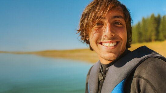 Lächelnder junger Mann in einem Neoprenanzug, vor einem ruhigen Gewässer und bewaldetem Ufer. Sonniger, klarer Himmel.