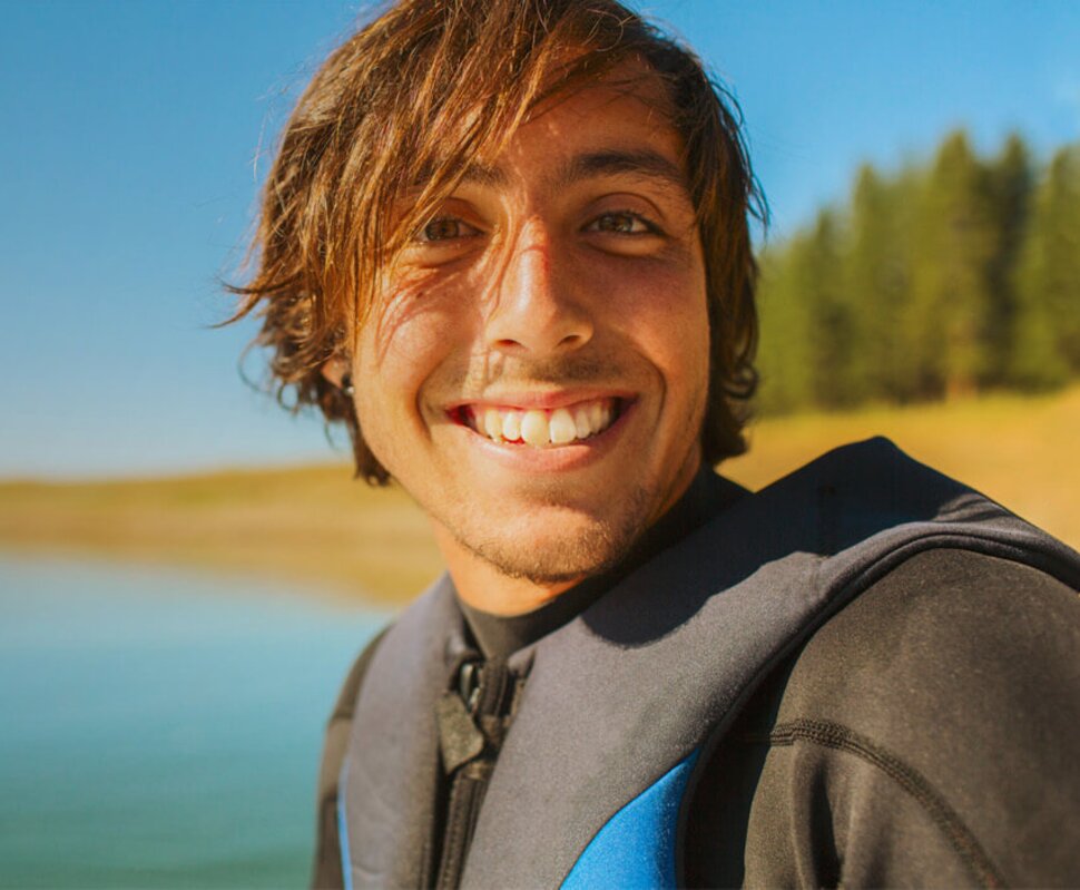 Lächelnder junger Mann in einem Neoprenanzug, vor einem ruhigen Gewässer und bewaldetem Ufer. Sonniger, klarer Himmel.