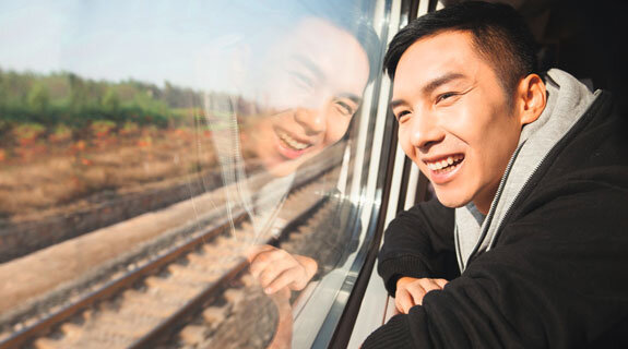 Junger Mann lächelt glücklich aus einem Zugfenster, mit Blick auf vorbeiziehende Landschaft und Schienen.