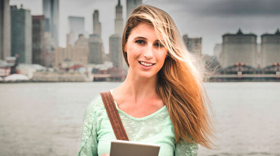 Junge Frau mit langen Haaren hält ein Tablet in der Hand, lächelt vor einer Stadtlandschaft am Wasser.