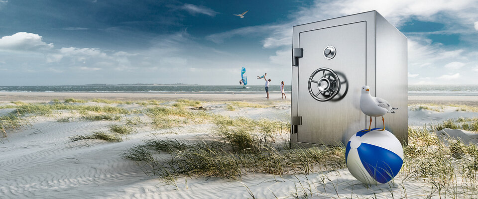 Ein großer Tresor steht am Strand, umgeben von Gras und Dünen, während eine Möwe auf einem Strandball sitzt.