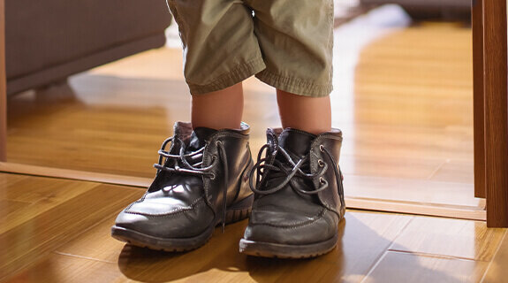 Ein Kind steht in zu großen, grauen Schuhen am Eingang eines Raumes auf einem Holzfußboden.