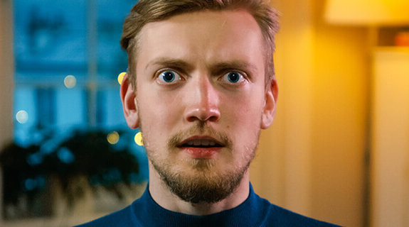 Ein junger Mann mit blonden Haaren und Bart schaut mit ernstem Gesichtsausdruck direkt in die Kamera, im Hintergrund unscharf.