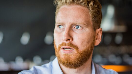 Ein Mann mit blondem Haar und rotbraunem Bart schaut überrascht. Im Hintergrund unscharfe, helle Lichtpunkte.