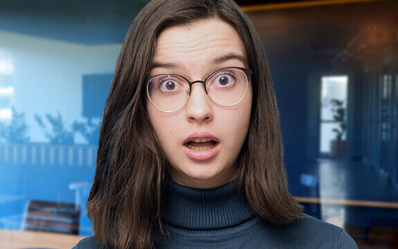 Junge Frau mit langen Haaren und Brille zeigt einen überraschten Gesichtsausdruck vor einem modernen Bürohintergrund.