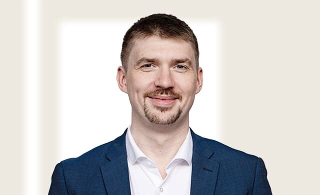 Männlicher Porträt mit kurzen Haaren, sportlichem Bart, blauer Jacke und weißem Hemd, lächelt vor neutralem Hintergrund.