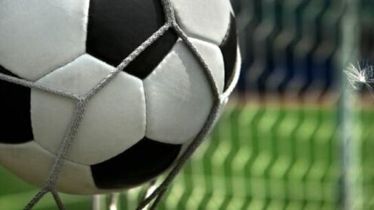 Ein Fußball rollt ins Netz eines Tores, mit einem verschwommenen Hintergrund auf einem grünen Spielfeld.