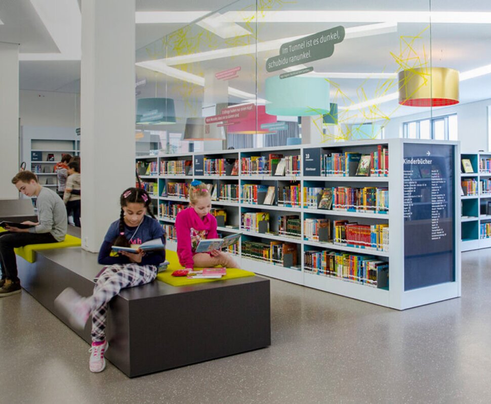 Moderne Bibliothek mit Lesebereichen, Regalen voller Bücher und mehreren Besuchern, die lesen oder lernen.