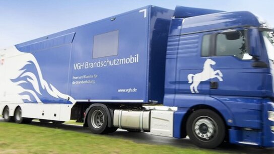 Blauer Lkw mit dem Aufdruck „VGH Brandschutzmobil“ fährt über eine Wiese, im Hintergrund ist der Himmel sichtbar.