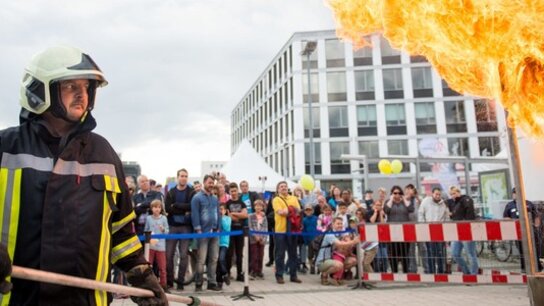 Feuerwehrmann mit Atemschutzgerät behandelt eine Flamme vor interessiertem Publikum auf einer Veranstaltung.