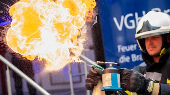 Ein Feuerwehrmann zündet mit einem Flämmchen eine große, gelbe Flamme an, während im Hintergrund ein Werbebanner sichtbar ist.