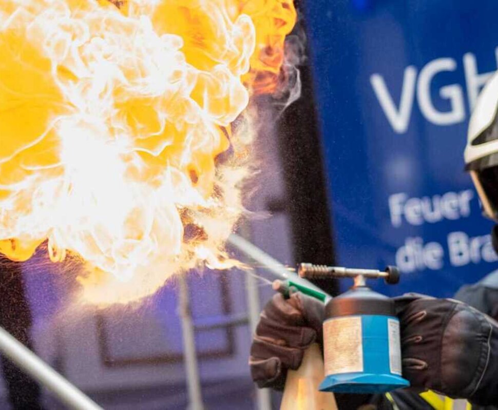 Ein Feuerwehrmann zündet mit einem Flämmchen eine große, gelbe Flamme an, während im Hintergrund ein Werbebanner sichtbar ist.