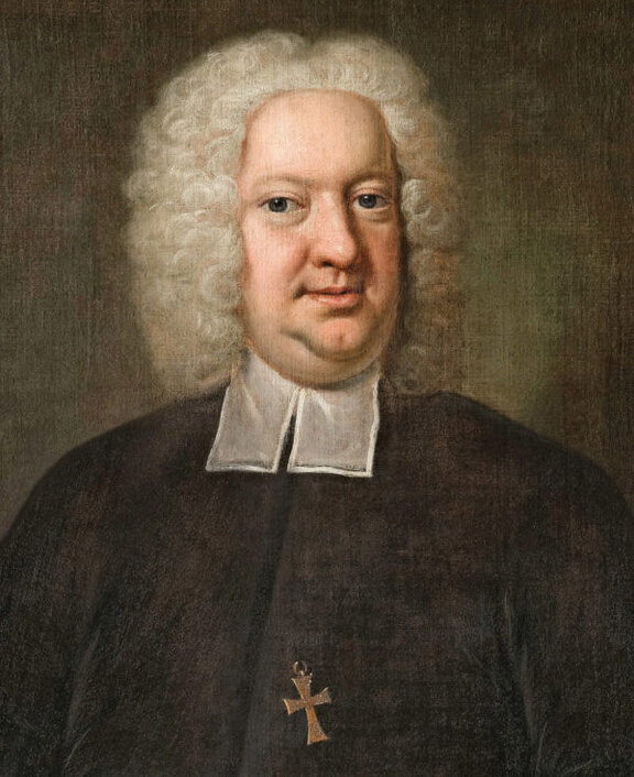 Porträt eines Mannes mit lockigem, weißem Haar, schwarzem Gewand und einem Kreuzanhänger um den Hals.