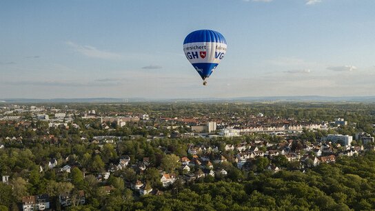 Heißluftballon schwebt über einer grünen Landschaft mit Häusern und städtischen Strukturen im Hintergrund.