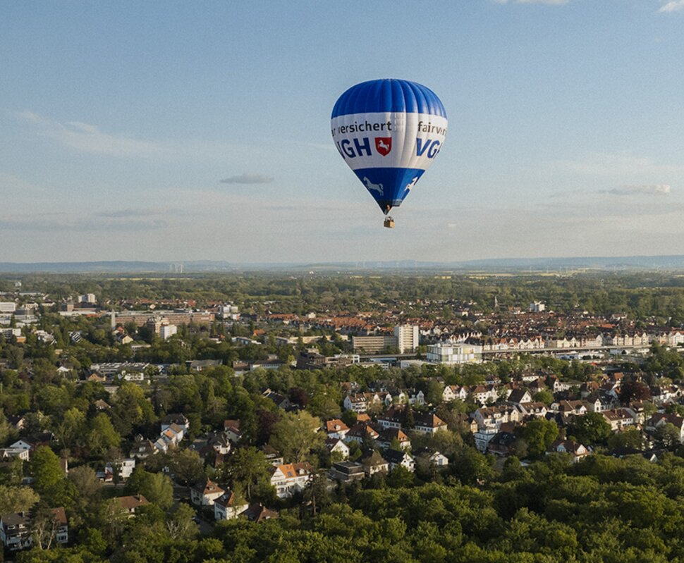 Heißluftballon schwebt über einer grünen Landschaft mit Häusern und städtischen Strukturen im Hintergrund.