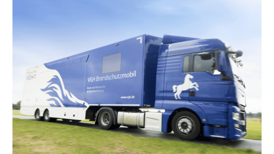 Blauer Lkw mit dem Schriftzug "VGH Brandschutzmobil" und einem weißen Pferdemotiv, fährt auf einer grünen Wiese.