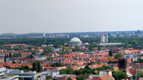 Panorama einer Stadt mit roten Dächern, grünen Bäumen und markanten Gebäuden wie einer Kuppel. Im Hintergrund ein Fernsehturm.