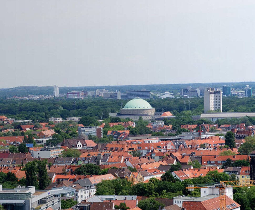 Panorama einer Stadt mit roten Dächern, grünen Bäumen und markanten Gebäuden wie einer Kuppel. Im Hintergrund ein Fernsehturm.