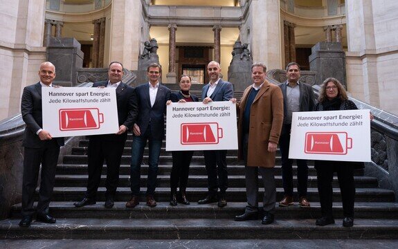 Gruppenzusammenkunft auf einer Treppe, die Plakate mit dem Slogan „Hannover spart Energie: Jede Kilowattstunde zählt“ zeigt.