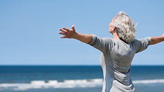 Eine ältere Frau mit grauen Haaren breitet die Arme weit aus und genießt den Blick auf das Meer und den blauen Himmel.