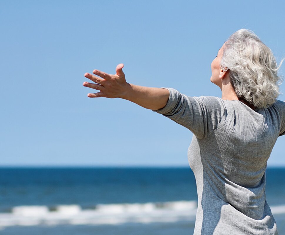Eine ältere Frau mit grauen Haaren breitet die Arme weit aus und genießt den Blick auf das Meer und den blauen Himmel.