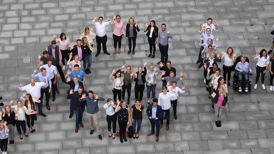 Gruppendynamik mit mehreren Personen, die fröhlich posieren und „NOW“ auf dem Boden bilden.