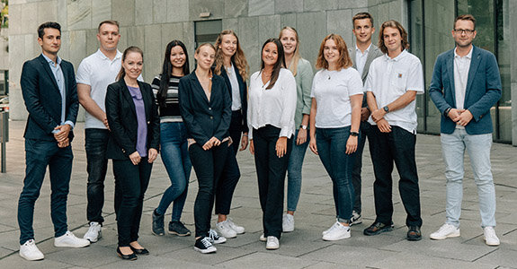 Gruppe junger Menschen in formeller und lässiger Kleidung posiert gemeinsam vor einer modernen Gebäude-Fassade.