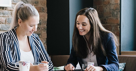 Zwei Frauen sitzen an einem Tisch, lächelnd und vertieft in eine Unterhaltung, umgeben von Backsteinwand und gemütlicher Atmosphäre.
