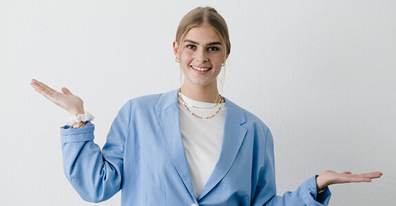 Junge Frau in einem blauen Blazer, lächelt und zeigt mit beiden Händen nach außen. Heller, neutraler Hintergrund.