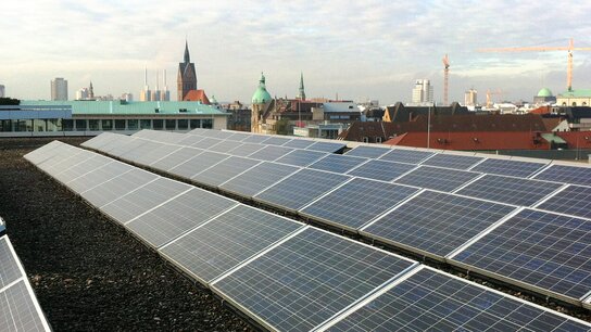 Solaranlage auf einem Dach mit Stadtansicht, darunter hohe Gebäude und Kirchtürme, im Hintergrund ein Kran.