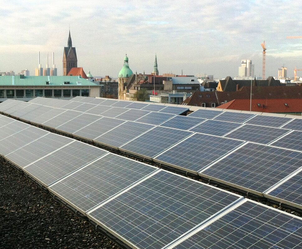 Solaranlage auf einem Dach mit Stadtansicht, darunter hohe Gebäude und Kirchtürme, im Hintergrund ein Kran.